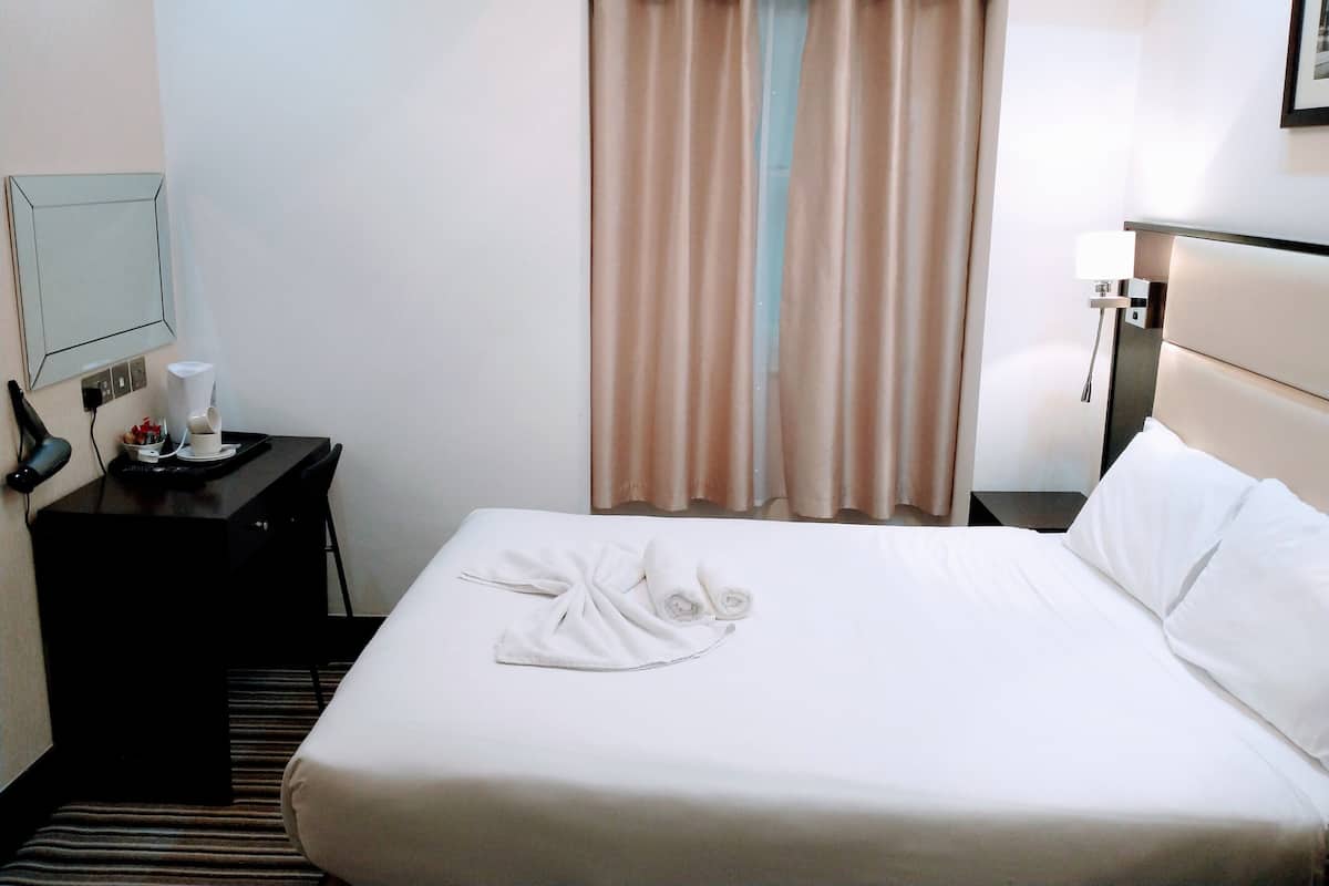 A hotel room with a neatly made bed, a bedside table with a lamp, a small table with a coffee maker, and a window with curtains.