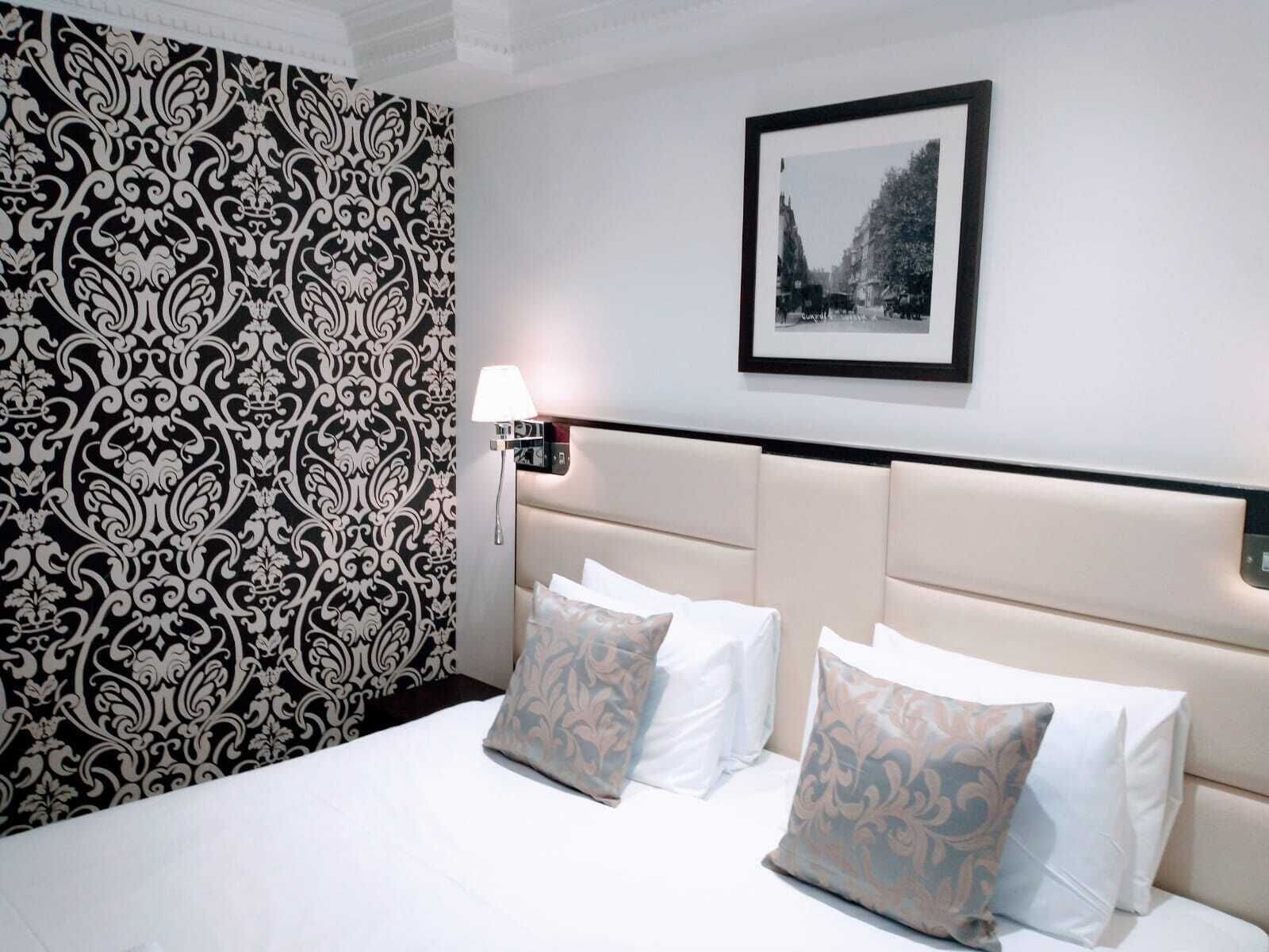 Ein Hotelzimmer mit strukturiertem Wandpapier, einem gerahmten Bild an der Wand, einer Nachttischlampe und einem Bett mit vielen Kissen.
