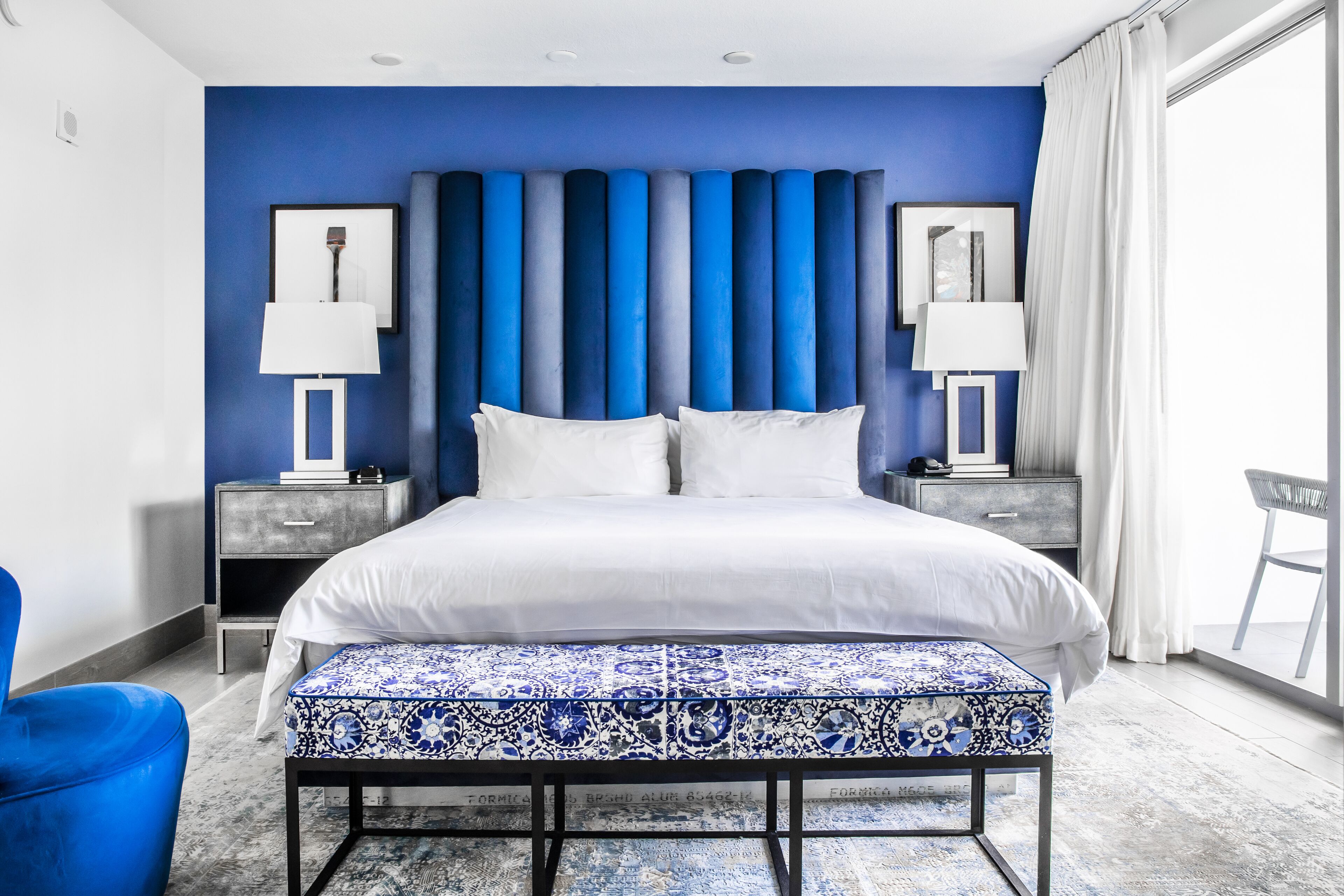 A modern bedroom with a blue accent wall, a large bed with white bedding, two bedside tables with lamps, and a blue patterned bench.