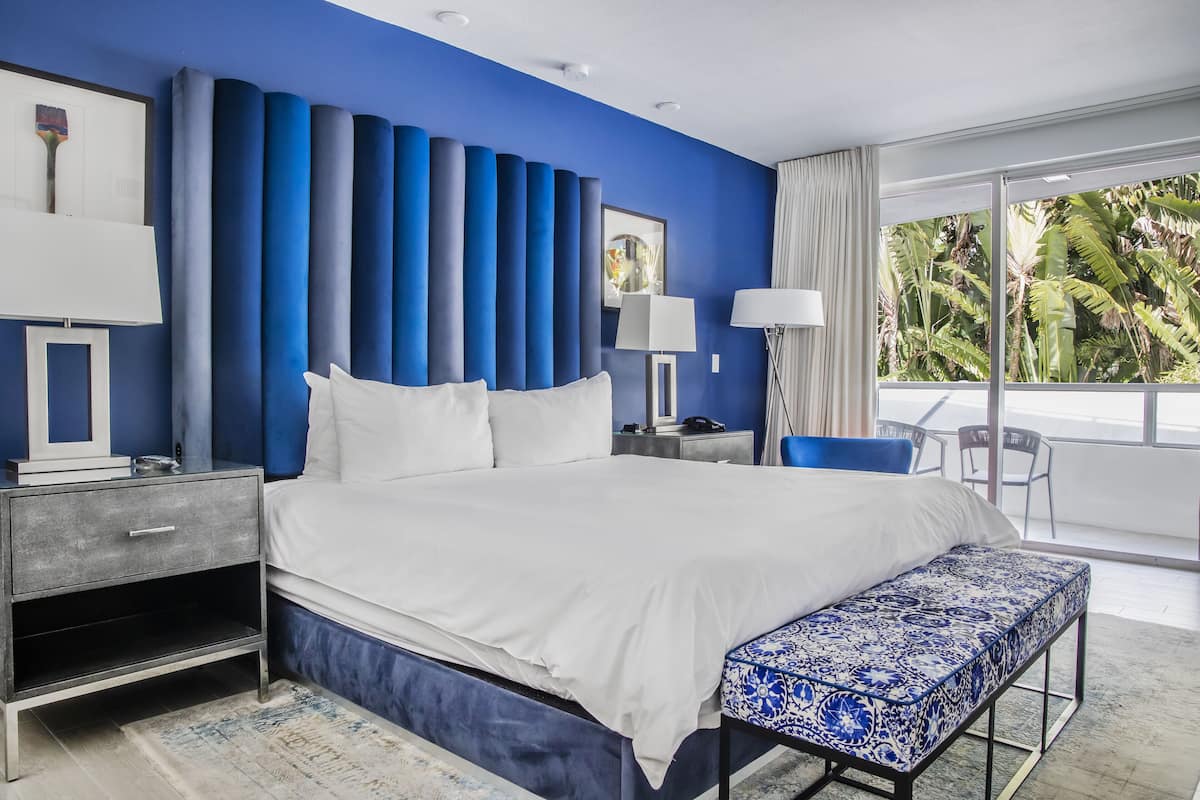 A modern hotel room with a large bed, a bedside table, a nightstand, a bench, and a view of the outdoors.