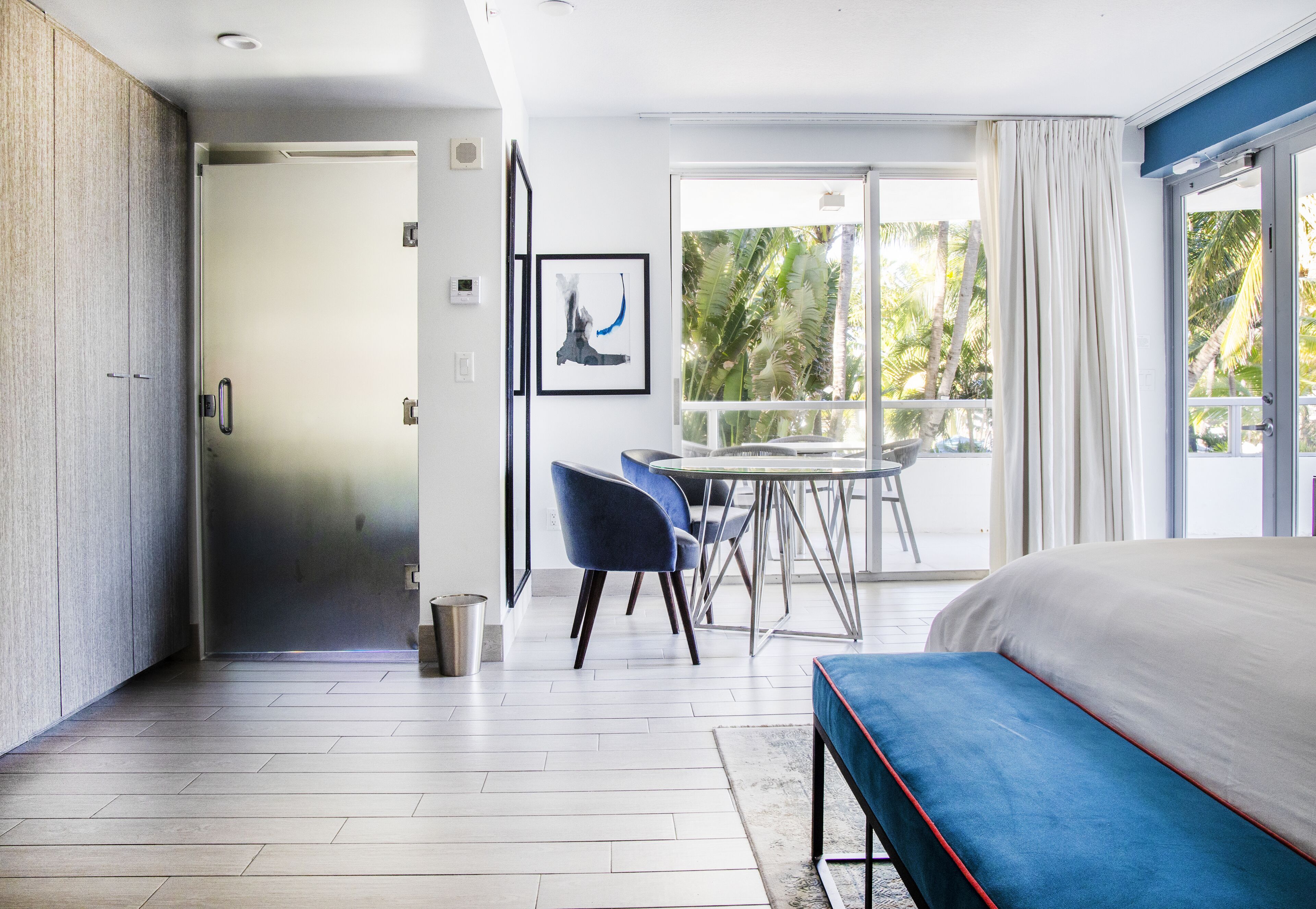 Une chambre d’hôtel moderne dotée d’un grand lit, d’un balcon offrant une vue sur les palmiers, d’un coin repas avec des chaises et d’une porte donnant sur une autre pièce.