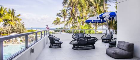 A rooftop lounge with wicker chairs and a pool view, surrounded by palm trees.