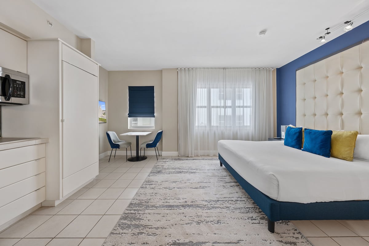 A modern hotel room with a bed, a desk, and a kitchenette.