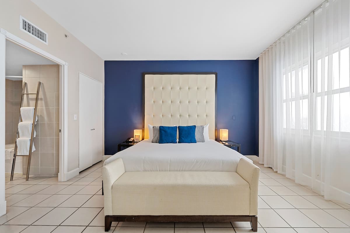 A spacious bedroom with a large bed, a nightstand, a bedside table, and a view of the outside through a window.
