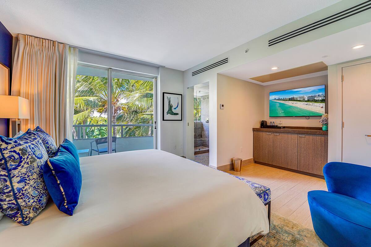 A hotel room with a large bed, a TV, a balcony with a view of palm trees, and a small kitchenette.