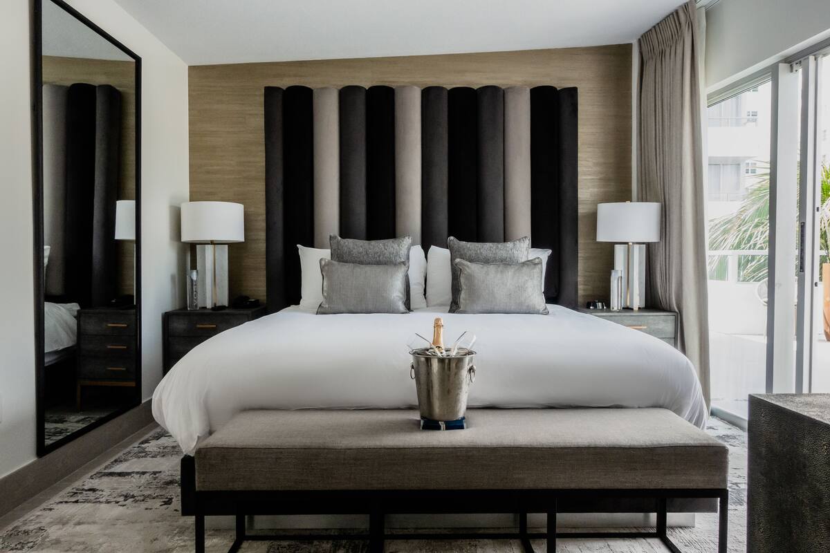 A modern hotel room with a large bed, a bedside table with a lamp, a nightstand, and a large mirror.