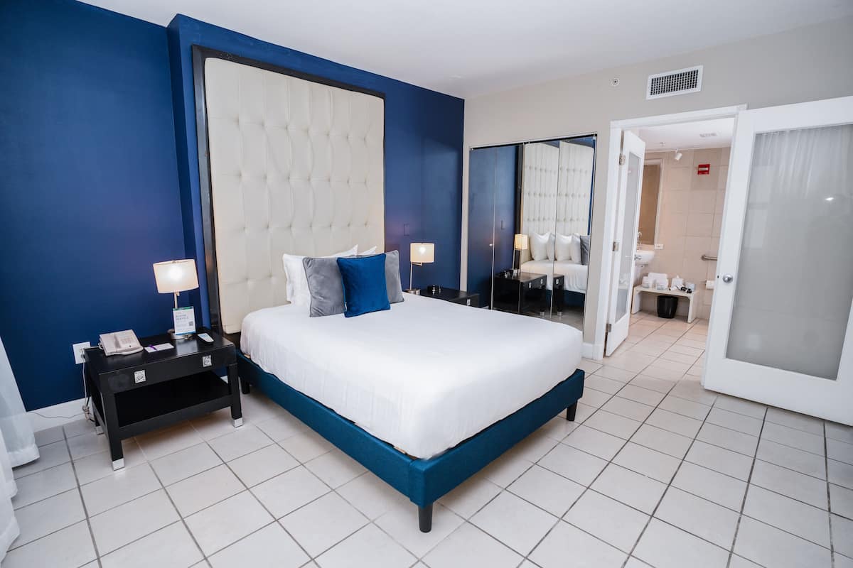A modern hotel room with a large bed, a nightstand, a lamp, and a bathroom visible through an open door.