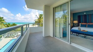 Ein Balkon mit Blick auf den Ozean und Palmen, ein Bett mit blauer Kopfteil und eine Glastür zum Zimmer.