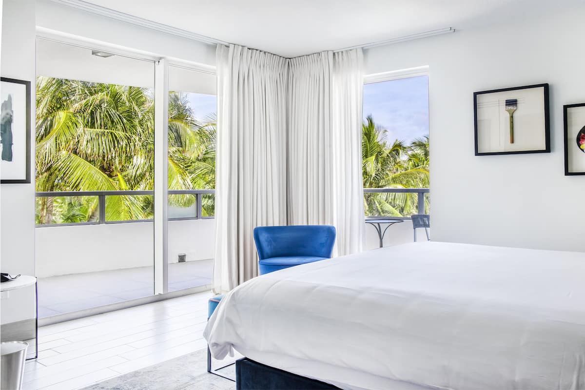 A bedroom with a large bed, a chair, a balcony with a view of palm trees, and framed artwork on the wall.