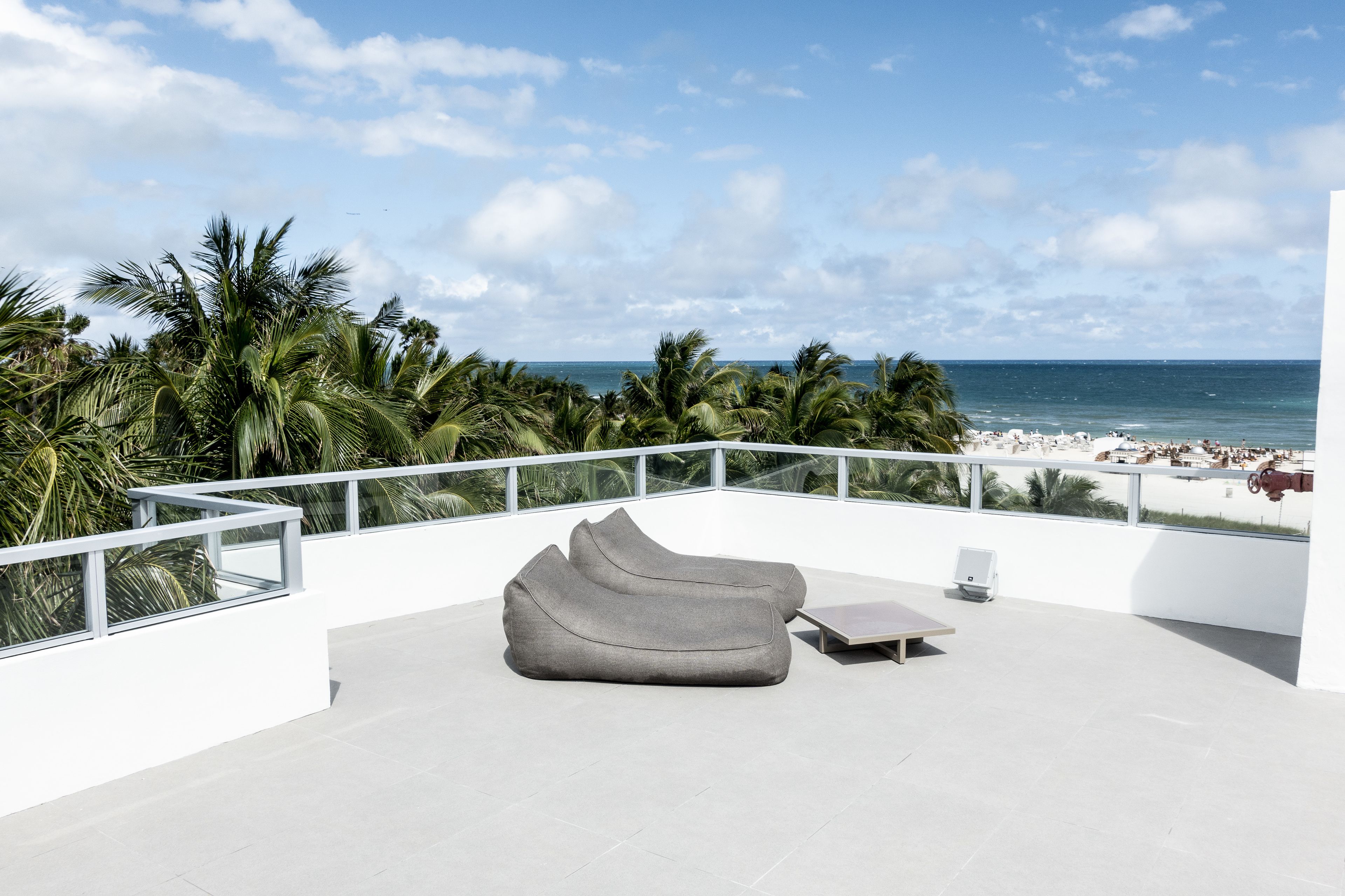 Ein moderner Balkon mit einer grauen Couch und einem kleinen Tisch, mit Blick auf einen Strand mit Palmen und dem Meer.