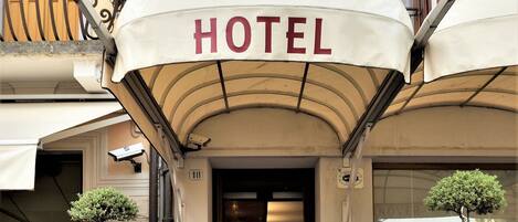 A hotel entrance with a red awning, potted plants, wicker furniture, and a sign indicating the hotel name and location.