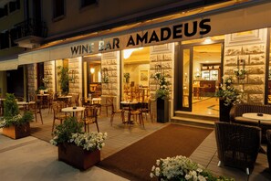 Un espacio al aire libre con mesas y sillas, la entrada de un bar de vinos, y plantas en macetas.