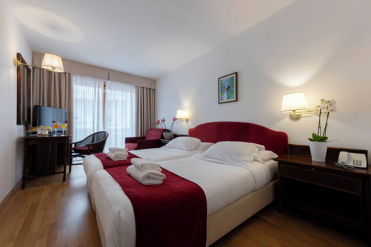 A hotel room with two beds, a desk with a TV, a chair, a red headboard, and two wall-mounted lamps.