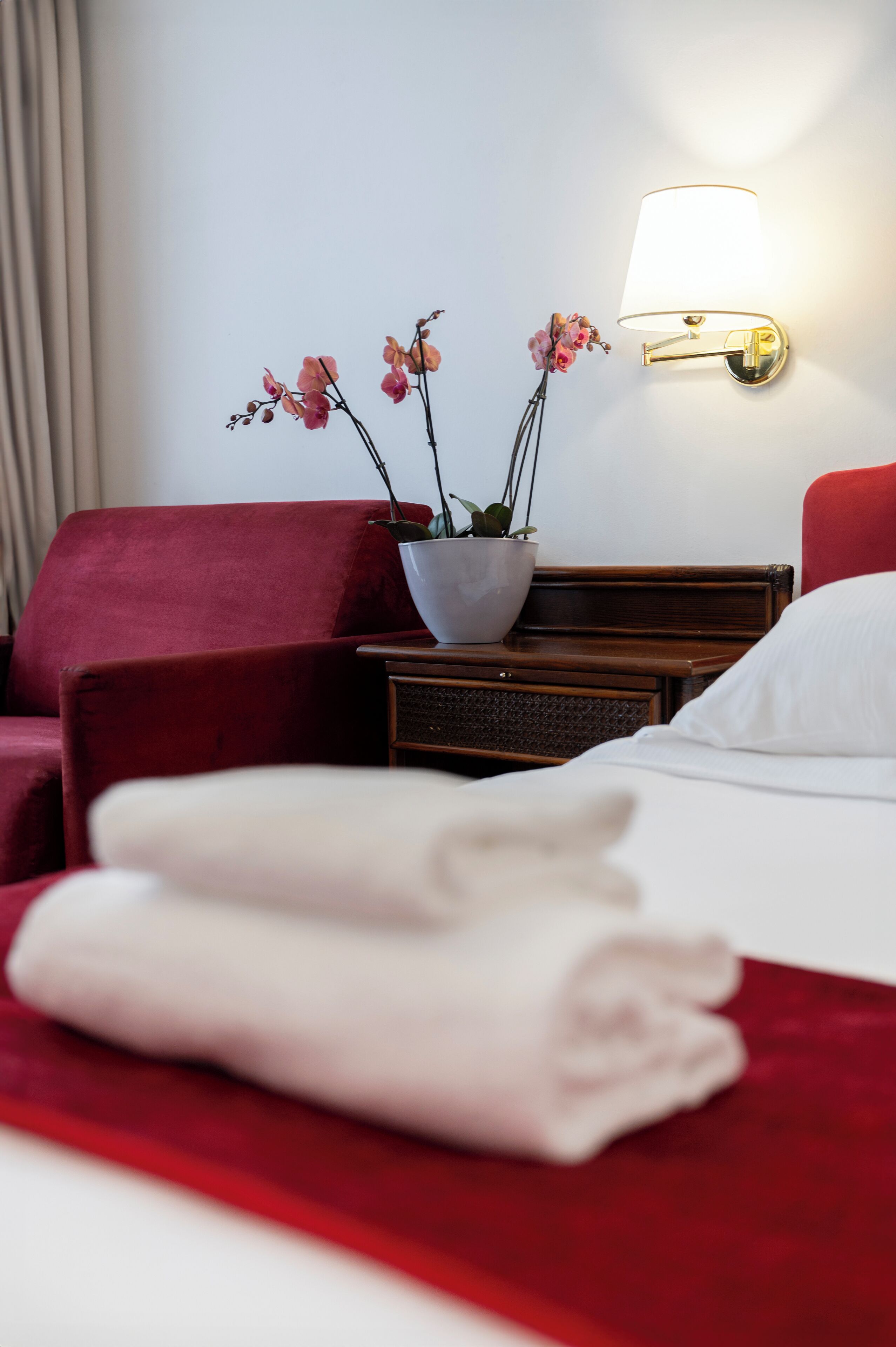A hotel room with a bed, a red armchair, a nightstand with a lamp, and a potted orchid plant.