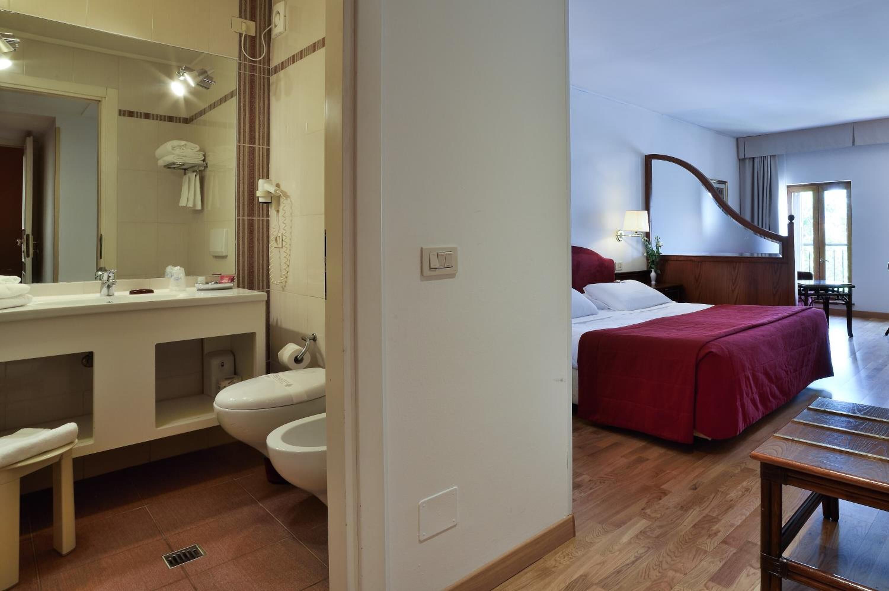 Habitación de hotel con piso de madera, cama con cubrecama rojo, cabecera de madera, mesita de noche, espejo, baño con lavamanos, inodoro y toallero.