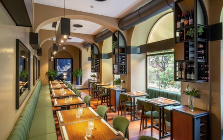 A restaurant with long wooden tables, green cushioned booths, and a wine rack on the wall.