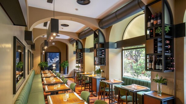 Un restaurante con largas mesas de madera, asientos tapizados en verde y una estantería para vinos en la pared.