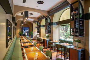 Un restaurante con largas mesas de madera, asientos tapizados en verde y una estantería para vinos en la pared.