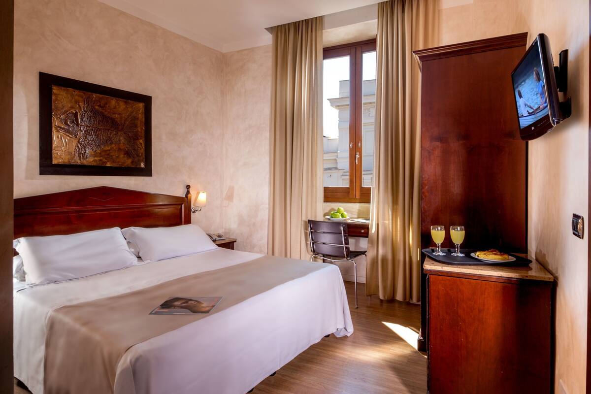 A hotel room with a large bed, a flat-screen TV mounted on the wall, a wooden desk with a chair, and a window with curtains.