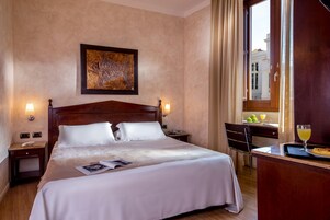 A hotel room with a large bed, a desk with a chair, a window with curtains, and a painting on the wall.