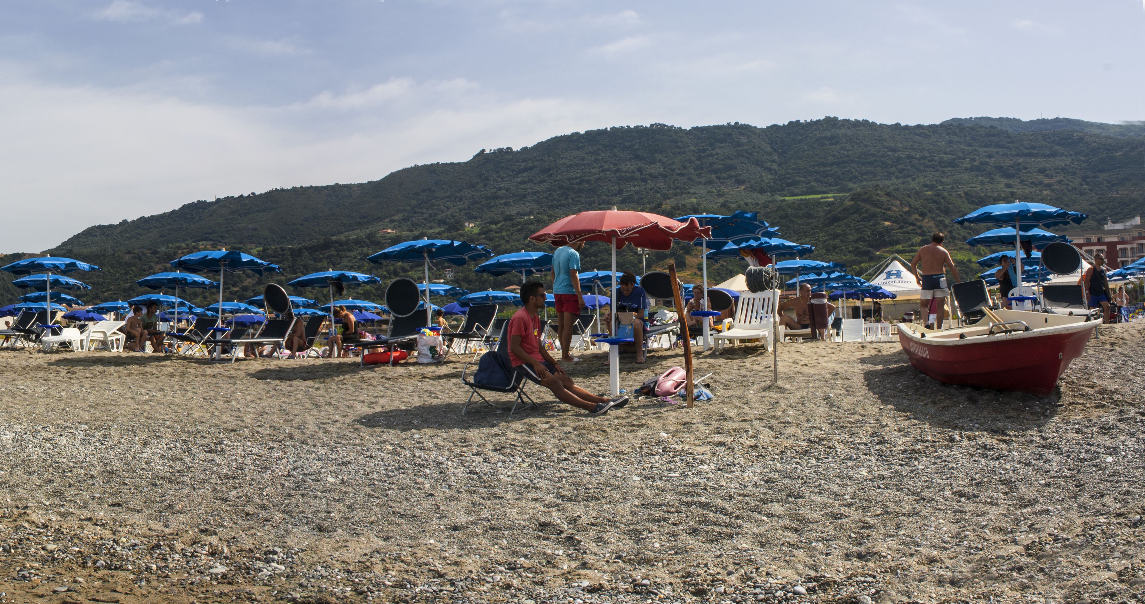 Una spiaggia con numerosi ombrelloni blu, una barca rossa e persone che si rilassano.