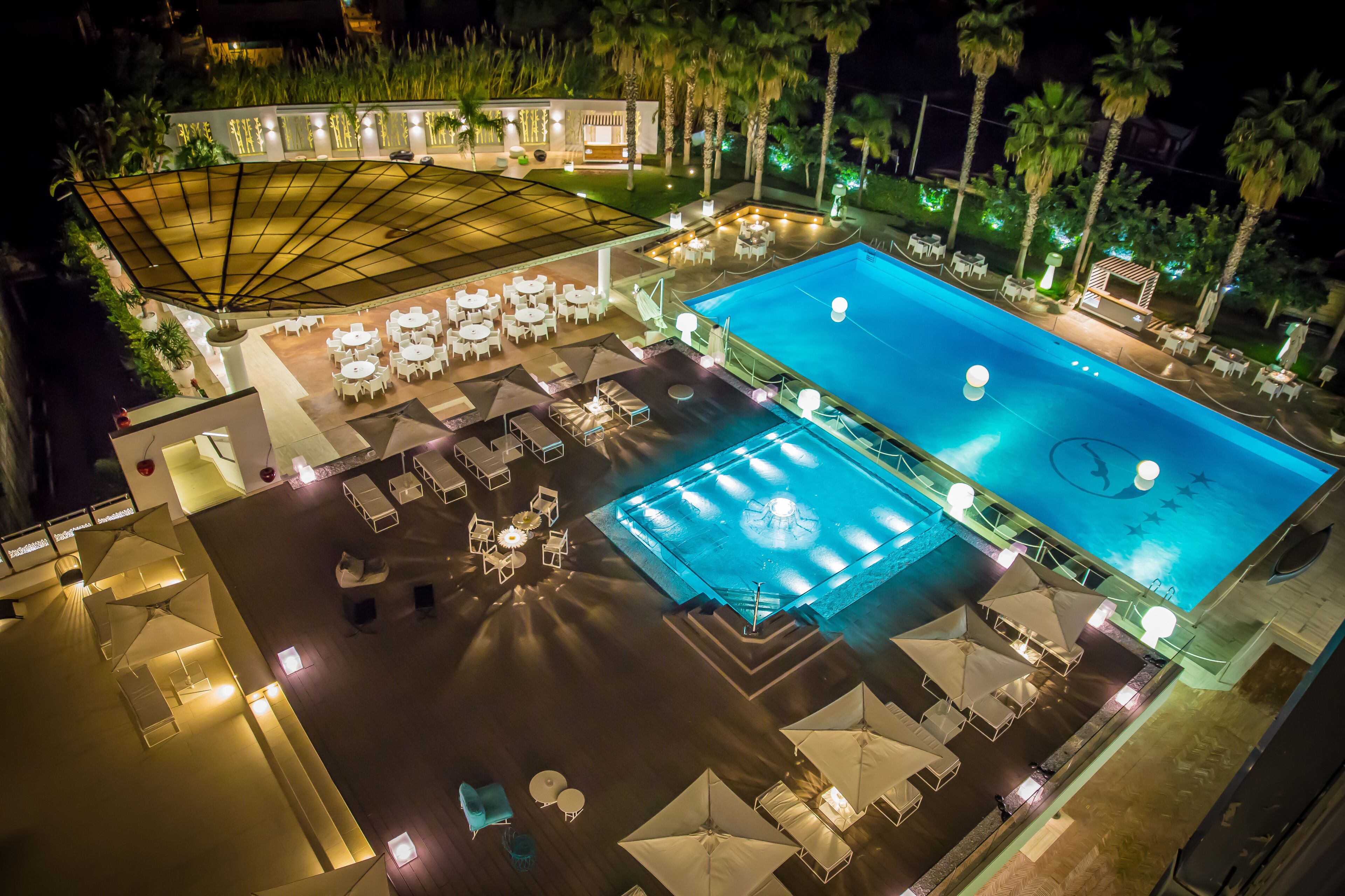 An outdoor swimming pool area with multiple lounge chairs and umbrellas, surrounded by palm trees and well-lit by ambient lighting.