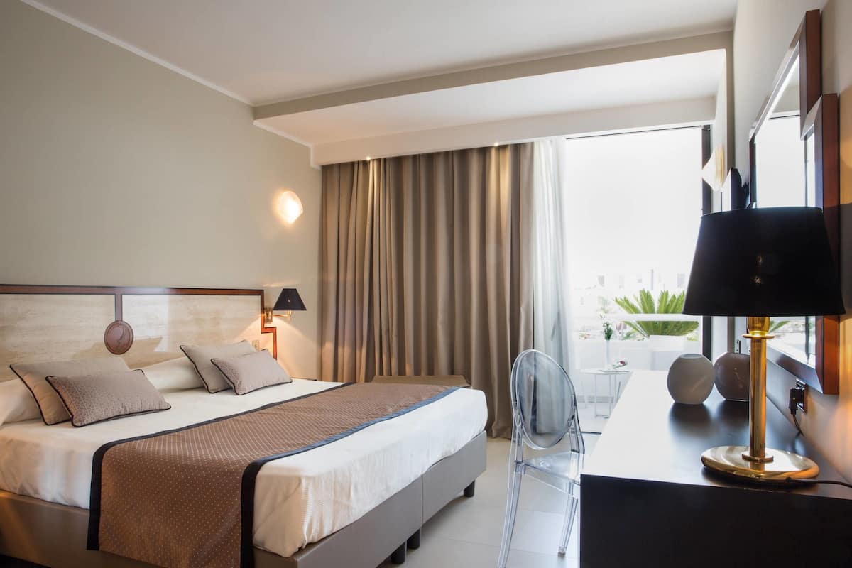 Una habitación de hotel moderna con una cama grande, un escritorio con lámpara y vistas a la ciudad a través de amplios ventanales.