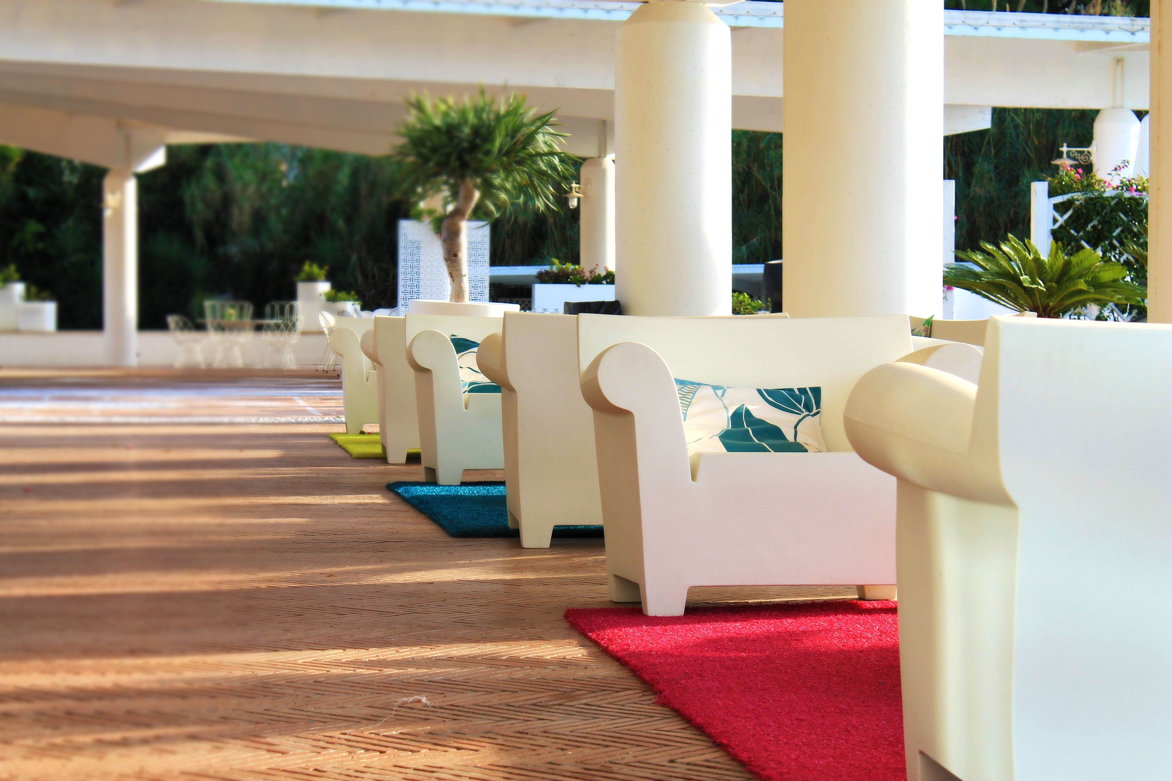 Una hilera de sillas blancas con cojines de colores, dispuestas sobre una terraza de madera con alfombra roja, bajo una estructura blanca con columnas.