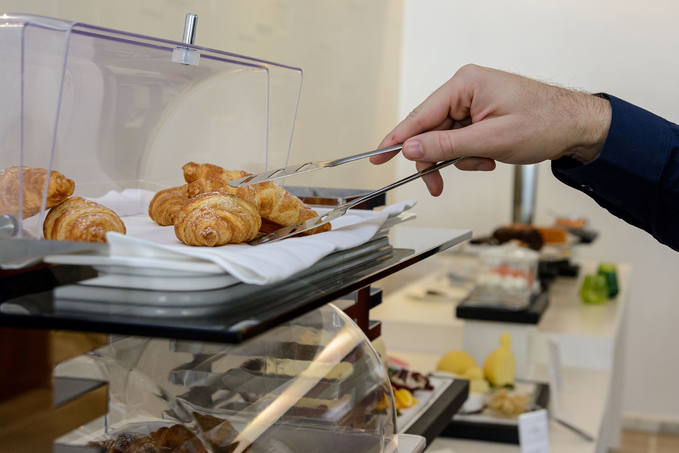 A hand using tongs to serve croissants from a glass display case.