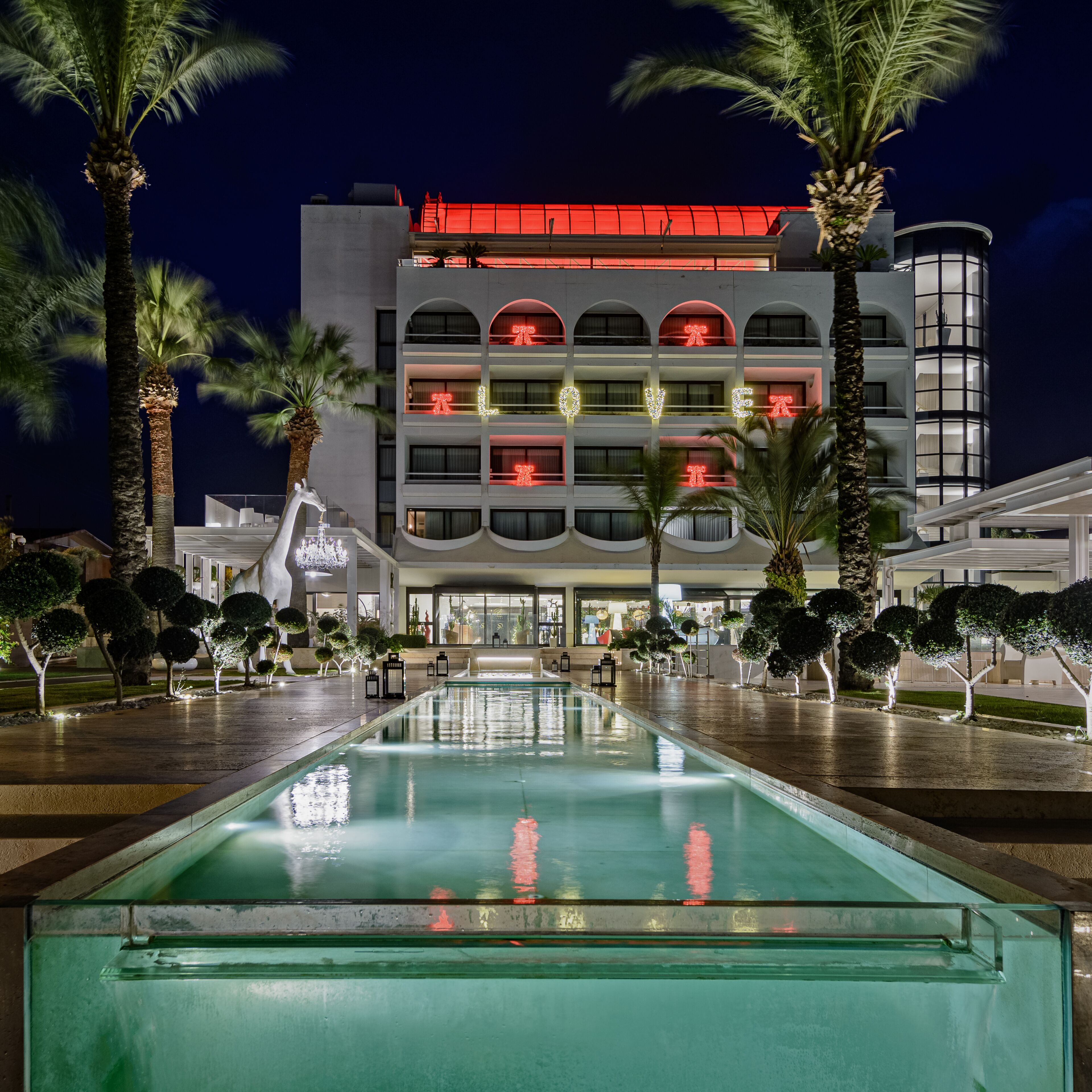 Un hotel con piscina, palmeras y un edificio moderno con ventanas iluminadas.