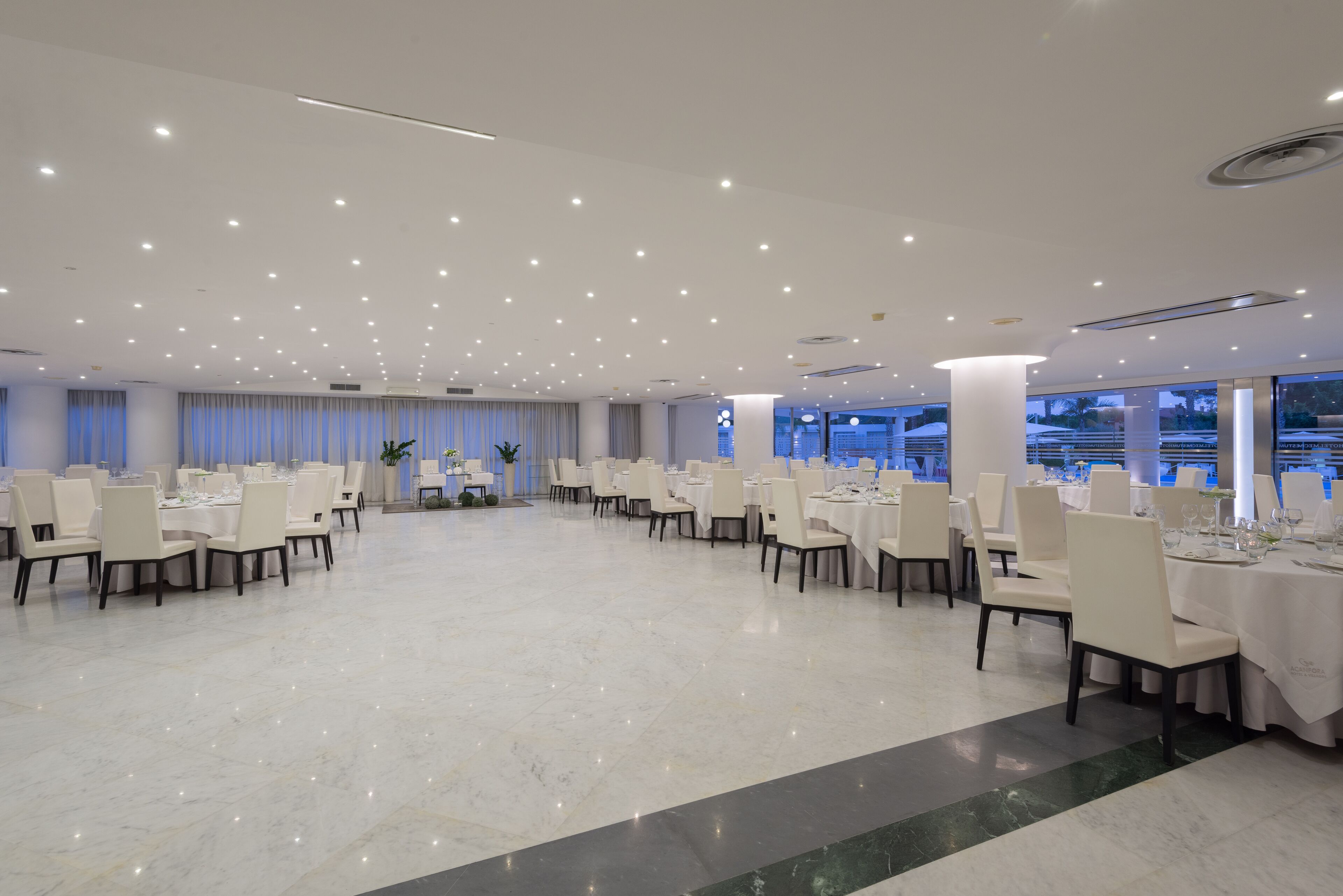 A spacious banquet hall with multiple round tables set for dining, large windows offering a view, and a polished marble floor.