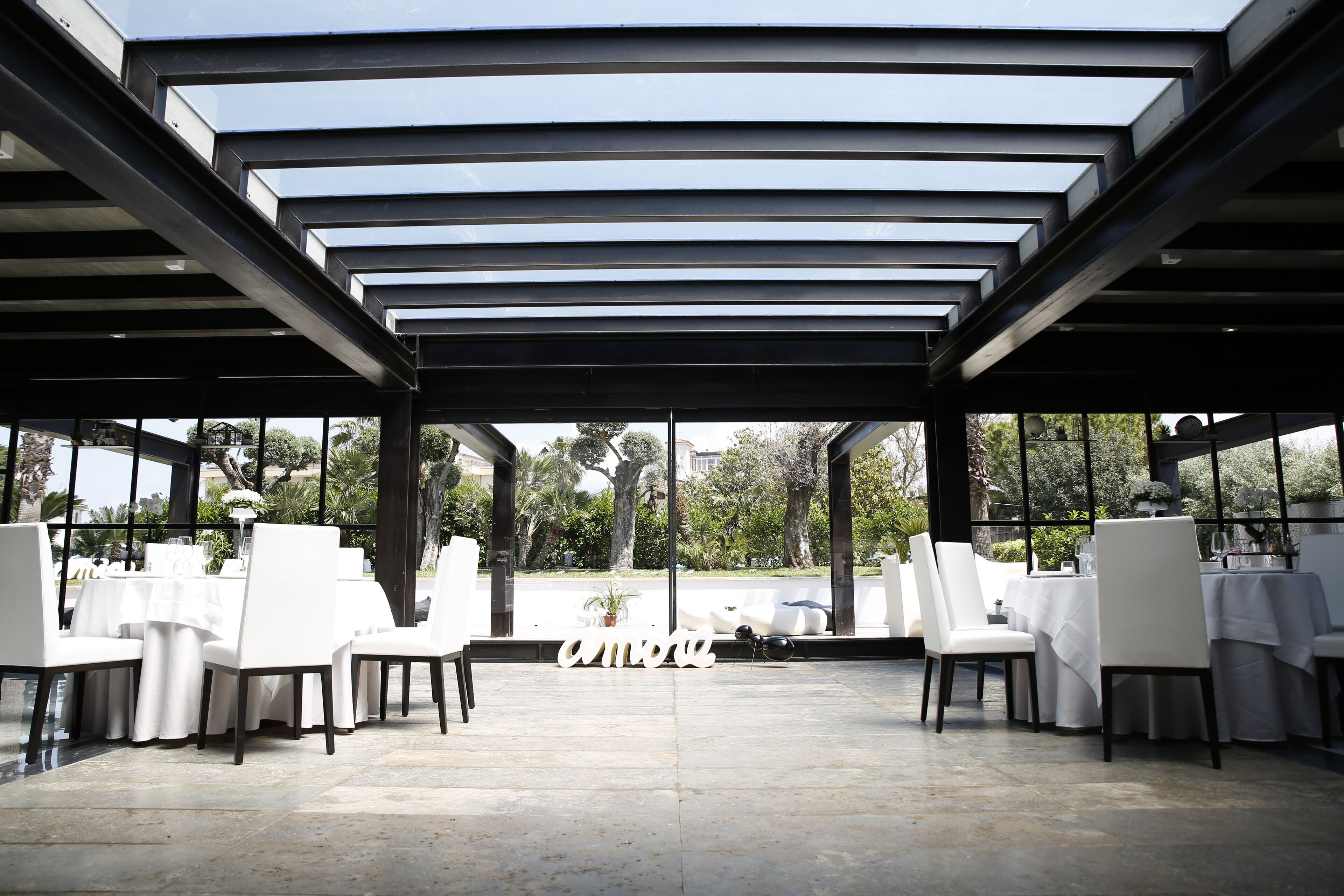 An outdoor dining area with white tables and chairs, a pergola structure, and a view of greenery.