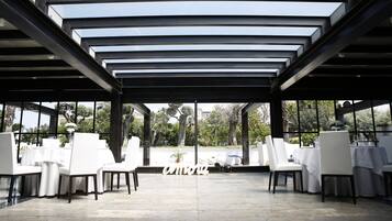 Un área de comedor al aire libre con mesas y sillas blancas, una estructura de pérgola y vistas a zonas verdes.