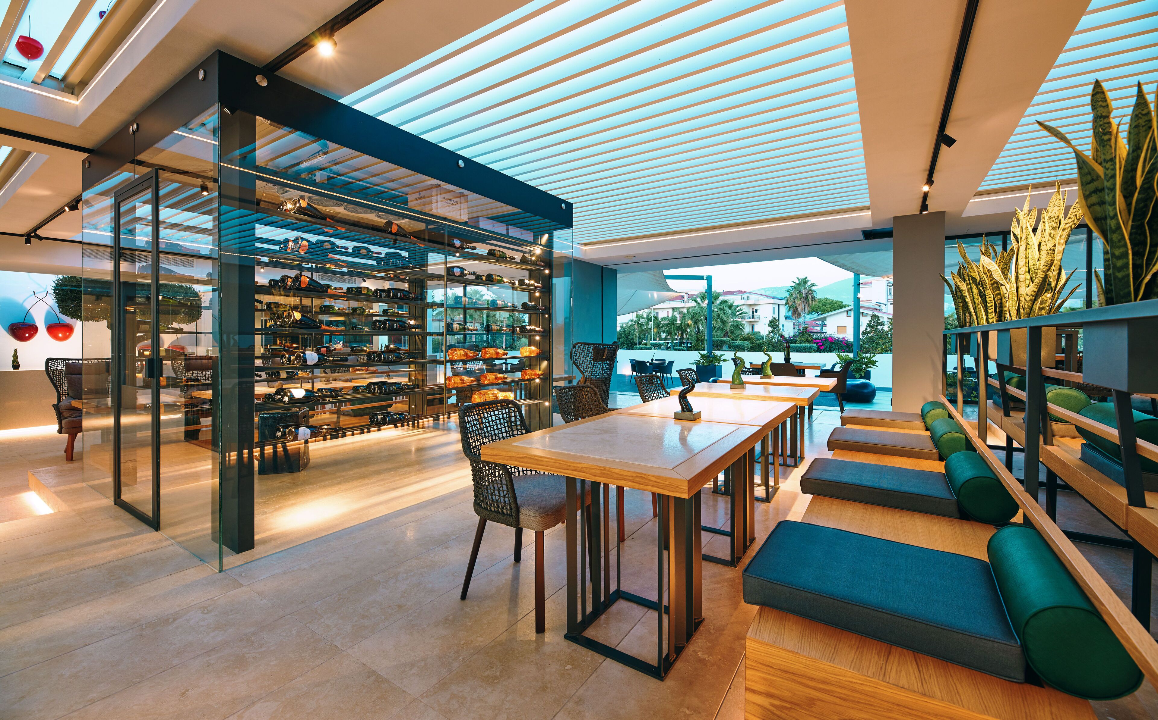 A modern interior with a glass display of wine bottles, wooden tables, and green seating.