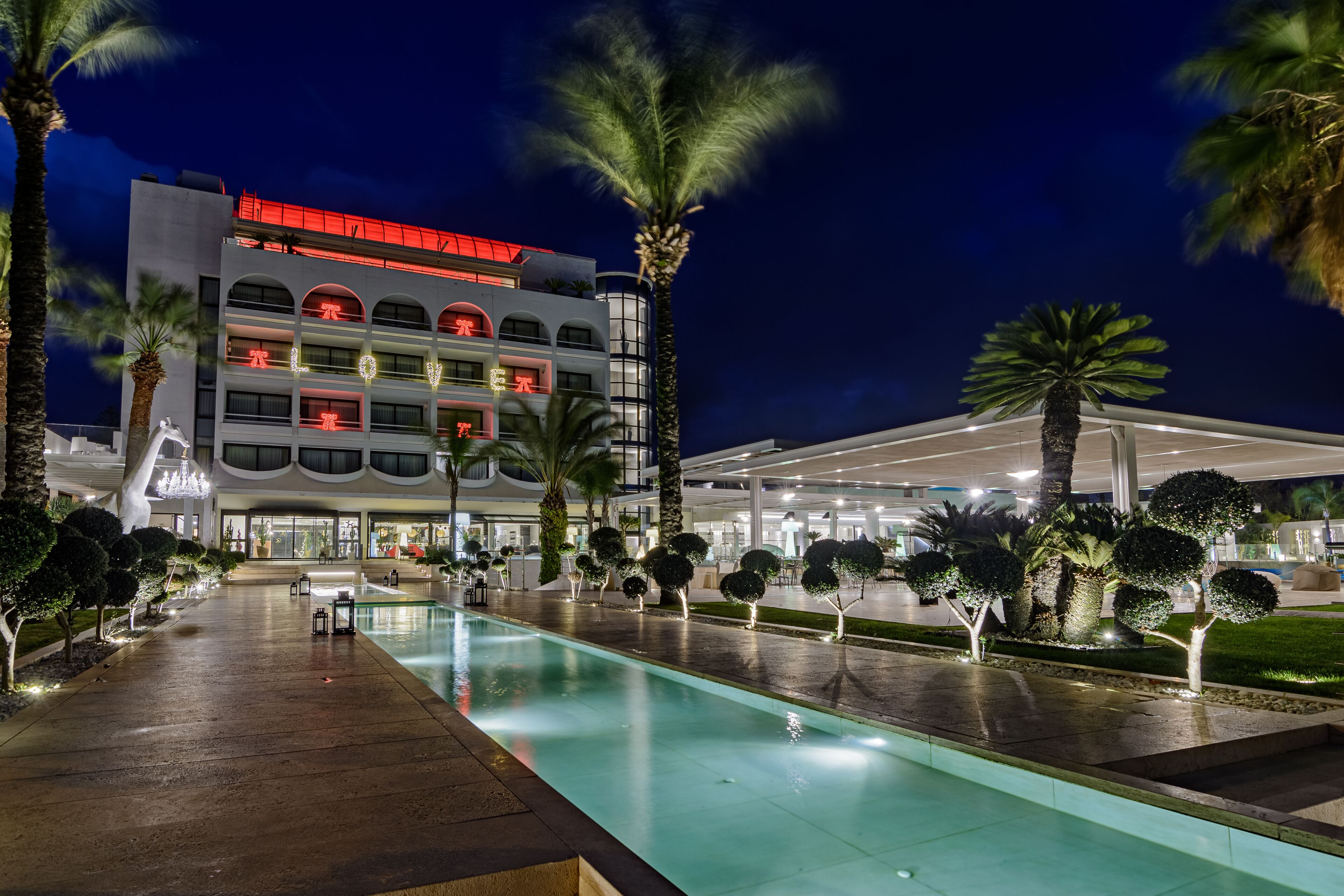 Een hotel met een zwembad, palmbomen en een modern gebouw met verlichte borden.