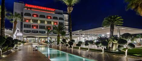 Un hôtel doté d’une piscine, de palmiers et d’un bâtiment moderne avec une enseigne lumineuse.