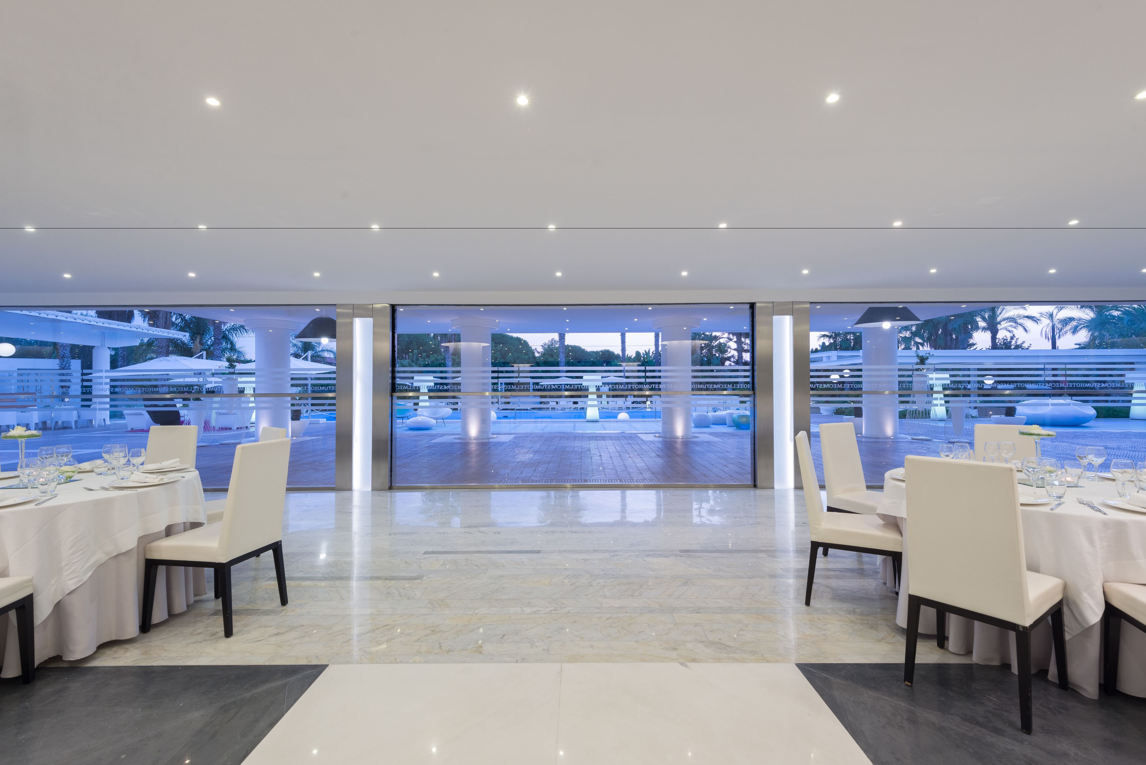 An indoor dining area with tables set for a meal, large windows, and a view of an outdoor pool area.