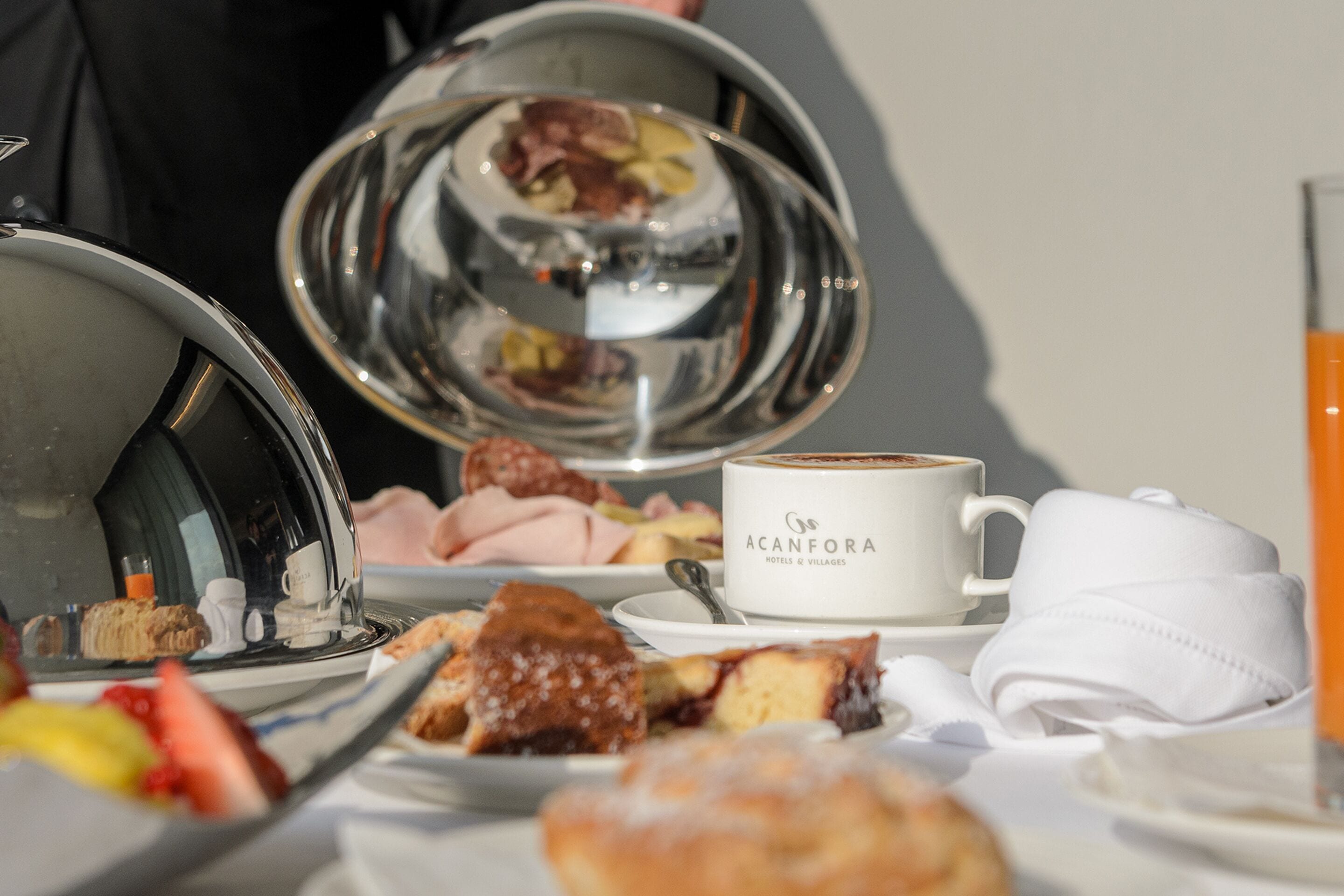 A breakfast setting with a coffee cup labeled "ACANFORA HOTELS & VILLAGES," a silver teapot, and various pastries on a tray.