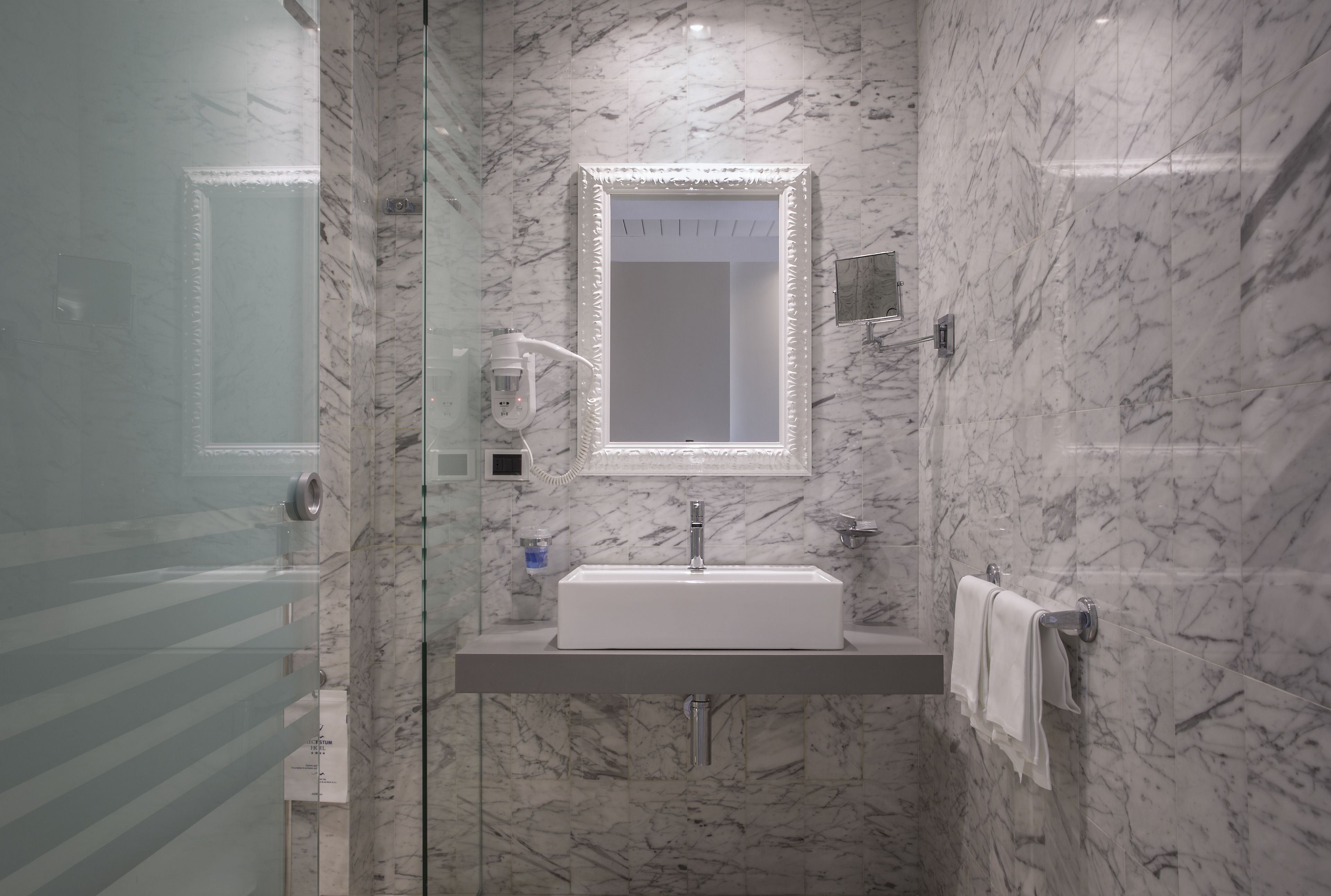 A modern bathroom with a marble wall, a rectangular mirror, a white sink, and a glass shower enclosure.