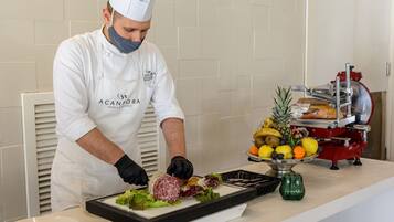 Uno chef con divisa bianca prepara cibo su un tagliere con verdure e carne, sullo sfondo si vedono un espositore di frutta e una affettatrice.