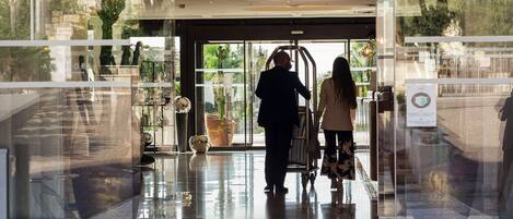 A modern hotel entrance with a glass facade, a reception desk, and a visible outdoor area with plants.
