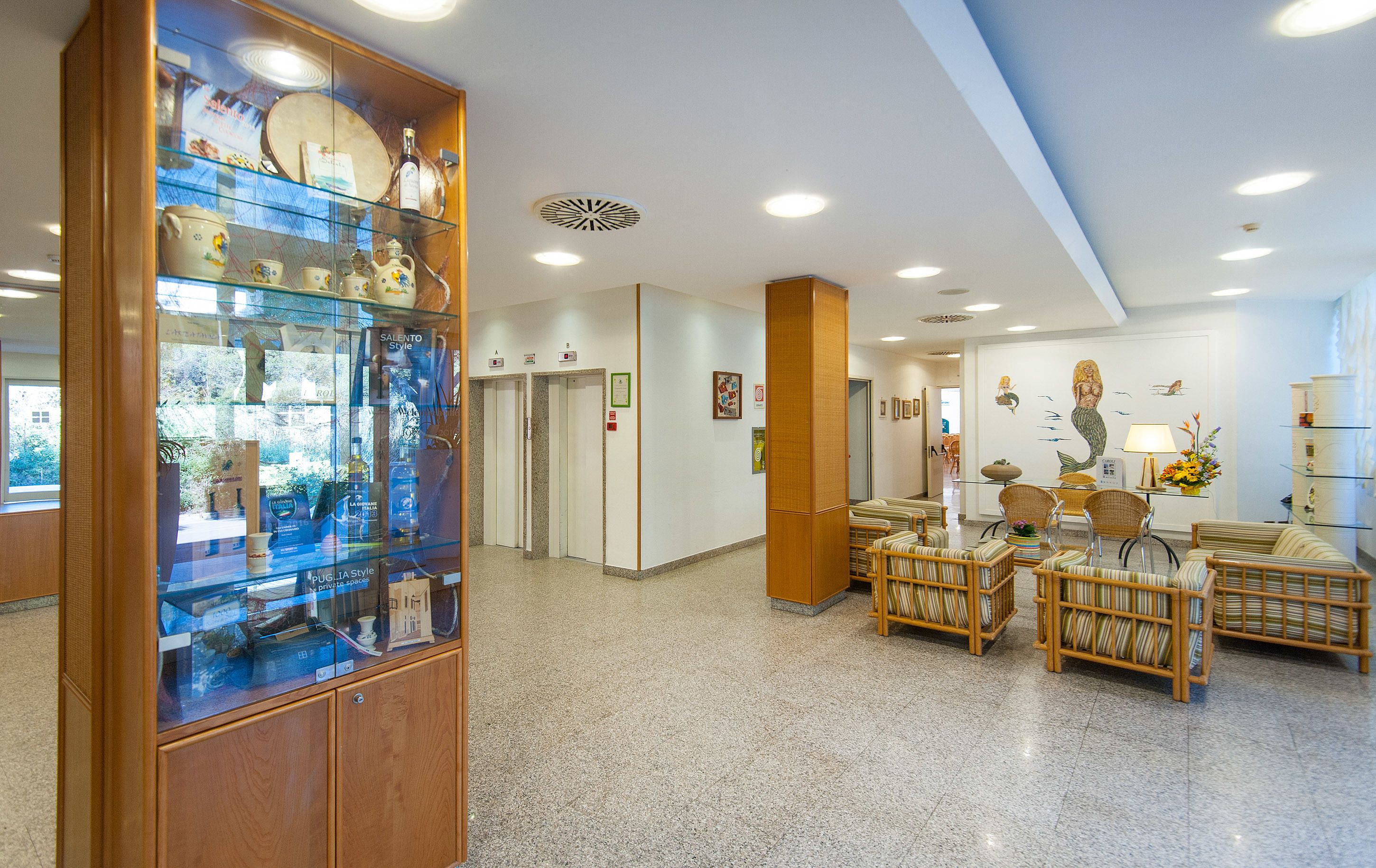 Vestíbulo de hotel con una vitrina de cristal, mobiliario de mimbre y un mural con sirenas.