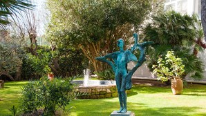 Una estatua de bronce en un jardín con una fuente, rodeada de exuberante follaje y un muro de piedra.