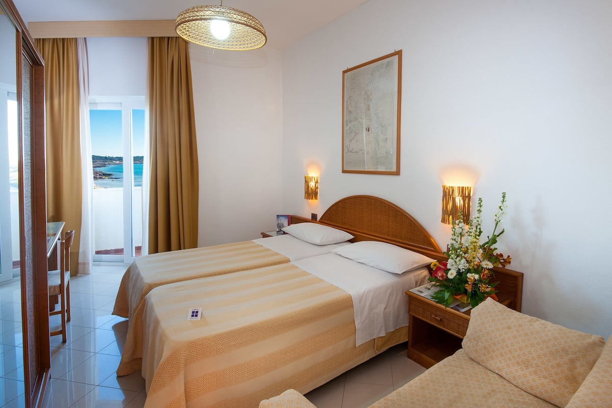 A hotel room with a bed, a nightstand, a lamp, a flower arrangement, and a view of the sea through the window.