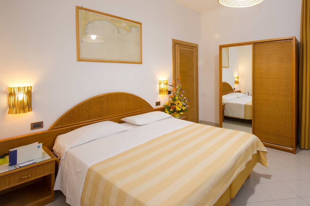 A hotel room with a bed, a nightstand, a mirror, a window with curtains, and a wall-mounted map.