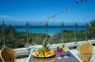 Una mesa con una bandeja de frutas y bebidas, con vista a una playa de aguas azules cristalinas y un faro a lo lejos.