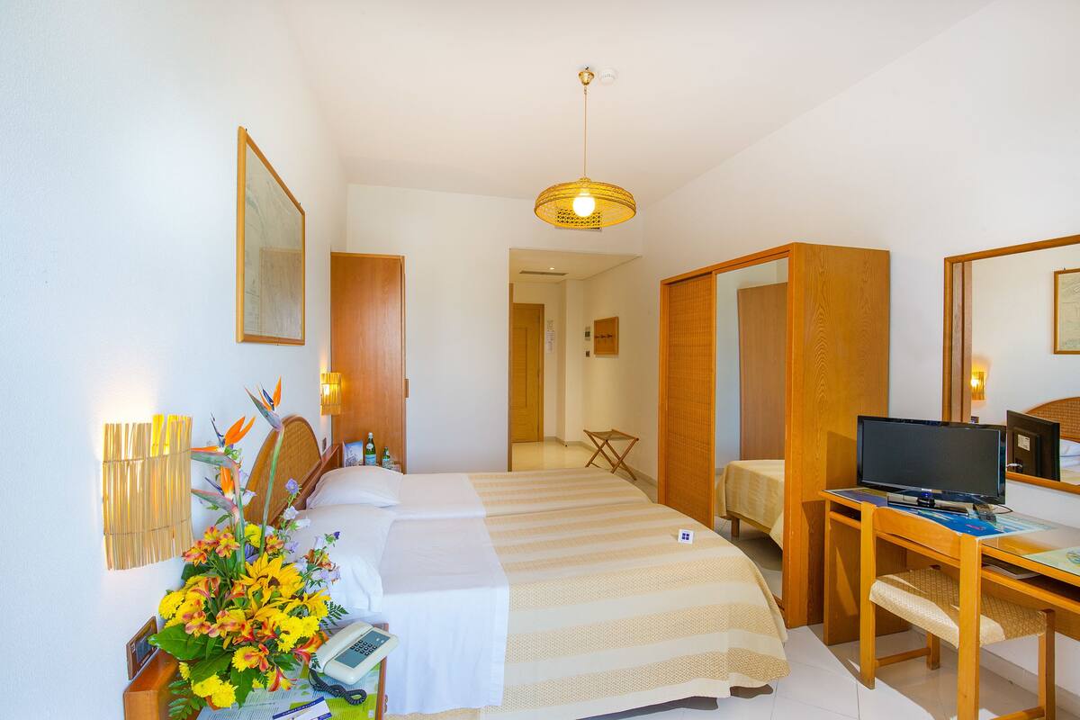 A hotel room with two beds, a desk with a computer, a small table, and a decorative plant.