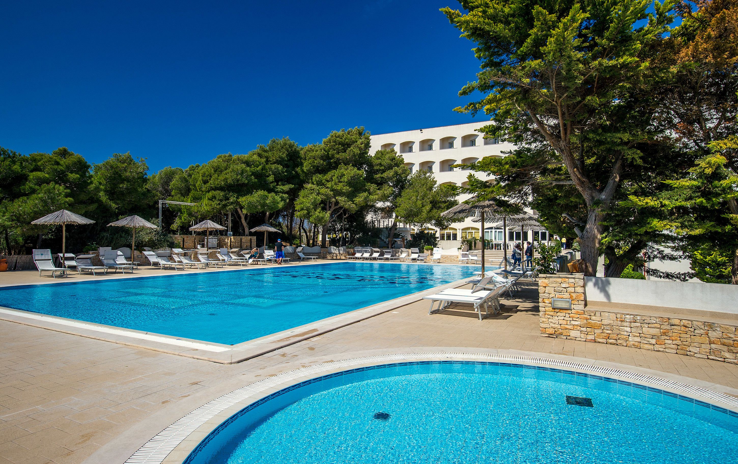 A large outdoor swimming pool with multiple lounge chairs and umbrellas, surrounded by trees and a building in the background.