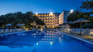 Una piscina con sillas de descanso y sombrillas, rodeada de árboles y un edificio con varios balcones.