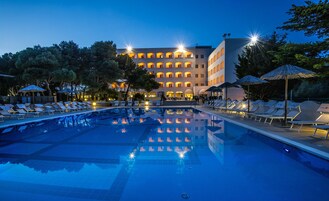 Una piscina con sillas de descanso y sombrillas, rodeada de árboles y un edificio con varios balcones.